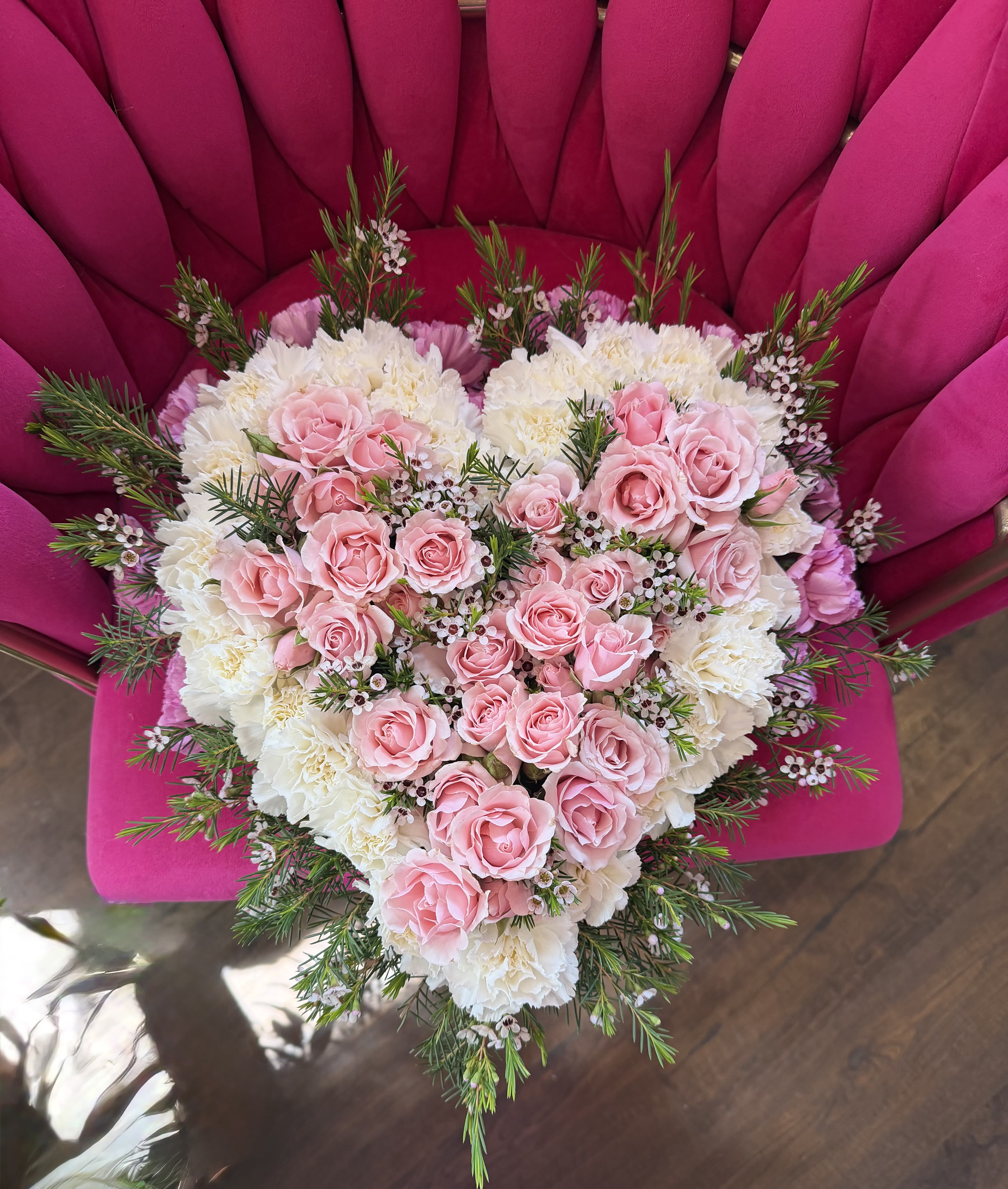 Eternal Heart - A heart-shaped floral arrangement of blush pink roses layered over creamy white carnations creates an immediate impression of tender devotion - ideal as an anniversary bouquet, a romantic gift, or a wedding tribute. The design reads as both affectionate and celebratory, the pink-and-white palette sending a clear message of love and warmth that will make the person you're thinking of feel truly cherished.  Clusters of soft pink roses form the dense center, set against a cushion of velvety white carnations for a pleasing contrast in color and texture. Fine sprigs of greenery and tiny white filler blossoms trace the heart's edges, adding an airy, lacy finish that keeps the composition light rather than heavy. The arrangement is presented on a vivid magenta chair in the display photo, which highlights the pastels and suggests an eye-catching way to present this piece at a party or ceremony.  Perfect for marking anniversaries, engagement celebrations, or as a romantic gesture, this arrangement communicates intimacy and care. Designed to be full and symmetrical, it holds its shape beautifully for display; for best longevity, keep it in a cool spot away from direct sunlight and replace water as needed if flowers are arranged in floral foam.