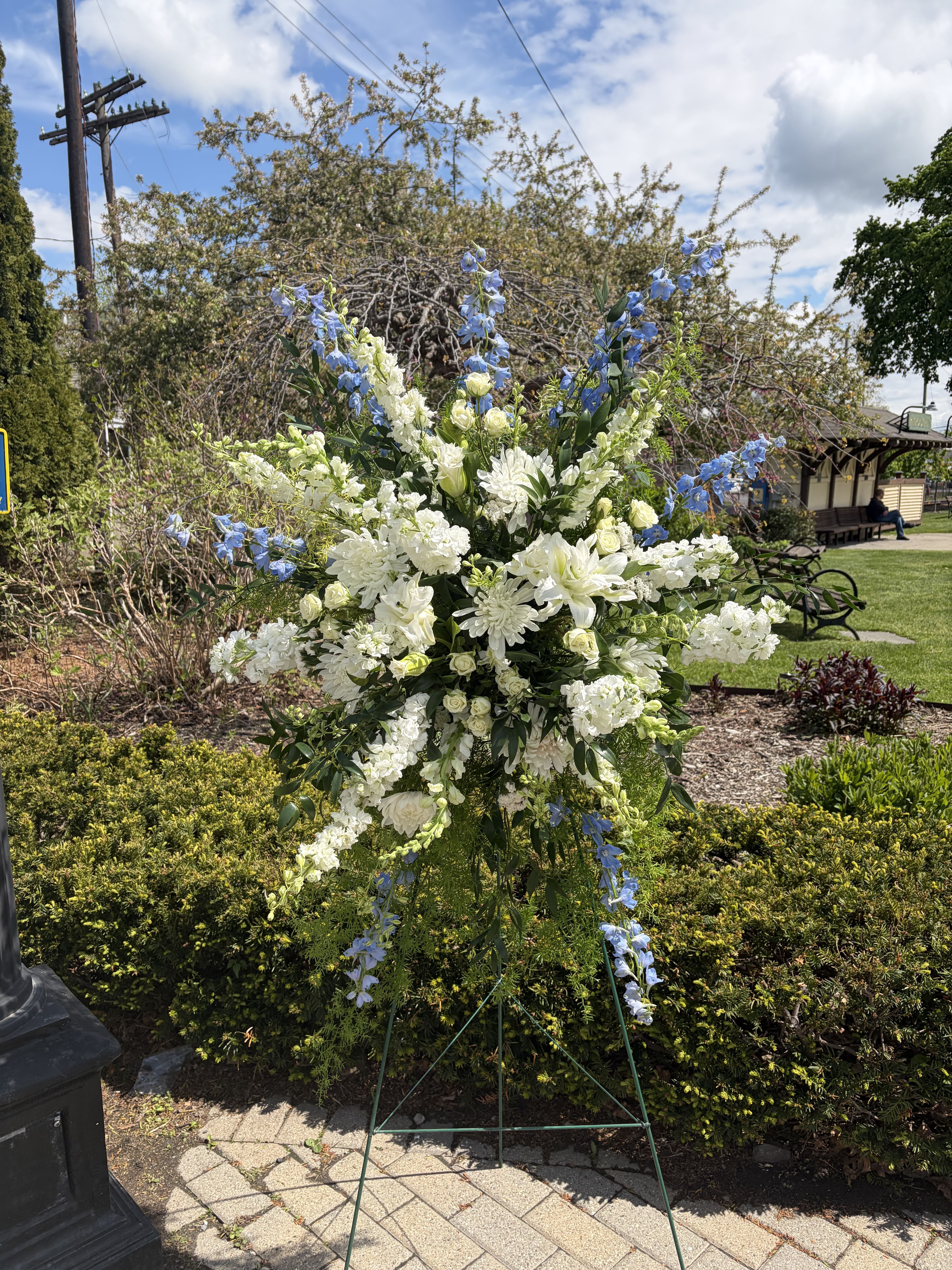 Double the Peace - A towering sympathy standing spray that speaks of calm and comfort, Double the Peace combines white and blue blooms to create a serene memorial display. Perfect for a funeral or graveside service, this flower arrangement places the ceremonial white center-a dense cluster of lilies, chrysanthemums, roses, and daisies-up front so the person you're honoring feels respectfully remembered, while tall blue snapdragons and other blue accents rise above and trail gently below for graceful vertical movement.  The composition pairs soft whites and cool blues with fresh green foliage for a formal, airy look. White lilies and roses lend classic fragrance and texture, chrysanthemums and daisies add fullness, and the blue snapdragons provide striking height and contrast. Sized as a large standing spray, it arrives ready to display on the included metal easel for immediate placement at the service or graveside.  Choose Double the Peace when you want to convey sympathy, reverence, and quiet support-the kind of tribute that helps the family feel seen in a difficult moment. For delivery to a funeral home or cemetery, please select the appropriate delivery option at checkout; keep blooms in a cool location away from direct sun until placed on the easel.