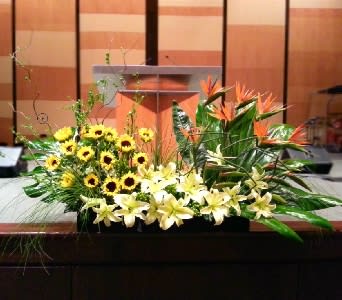 Long stage arrangement of yellow lilies, sunflowers, and orange tropical flowers in a low container