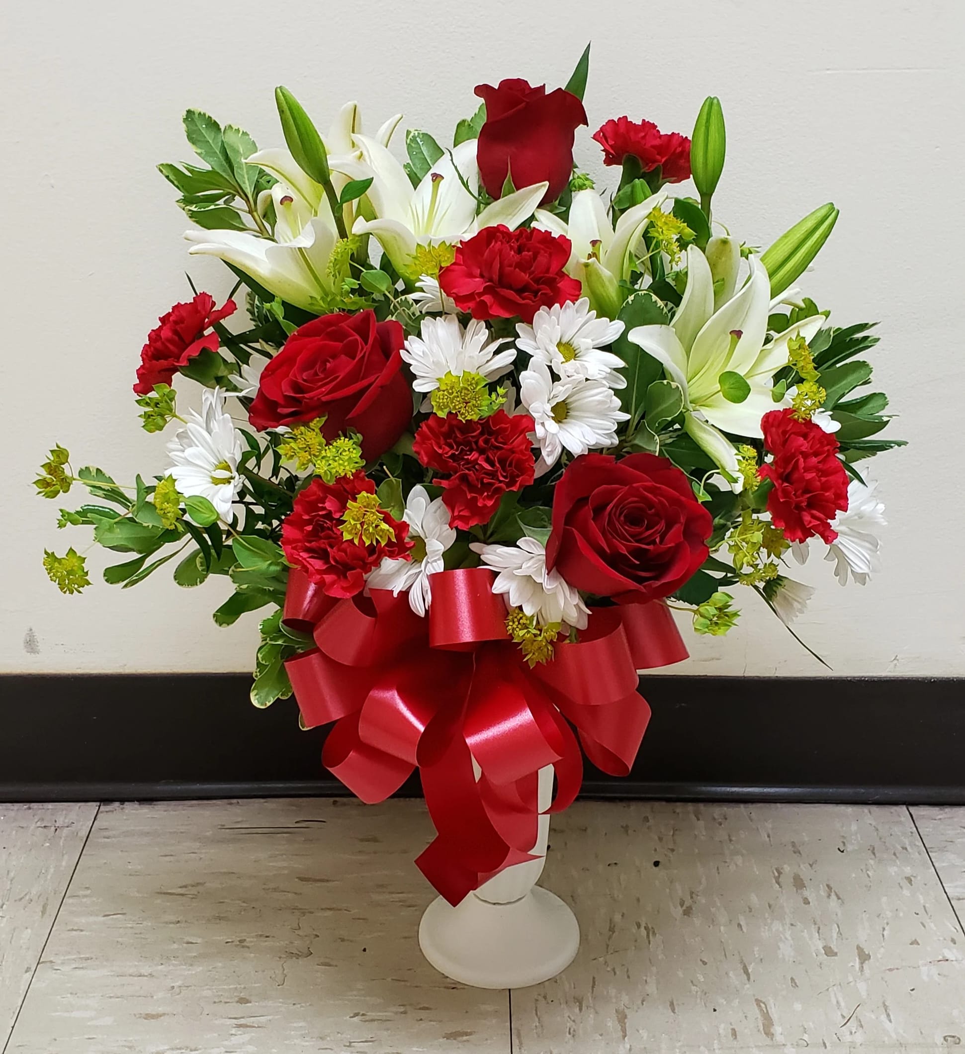Red roses, white lilies, and daisies arranged in a white vase with a red ribbon.