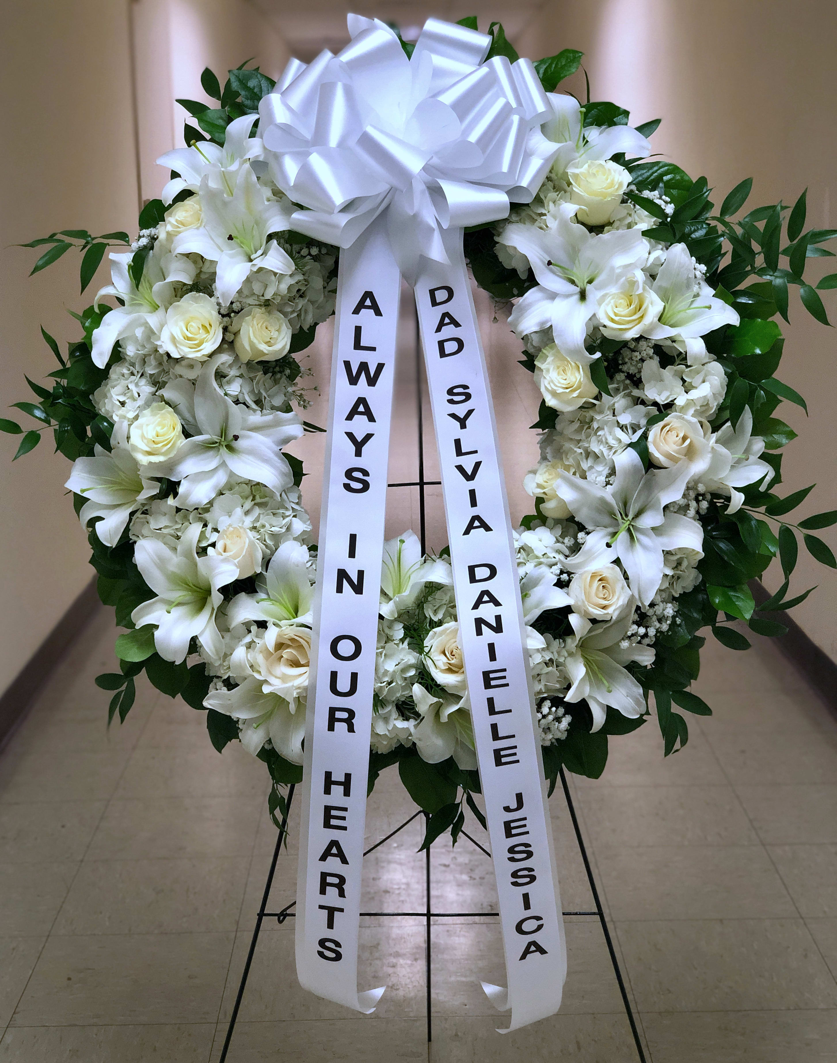 White funeral wreath with lilies, roses, and a large bow on a stand