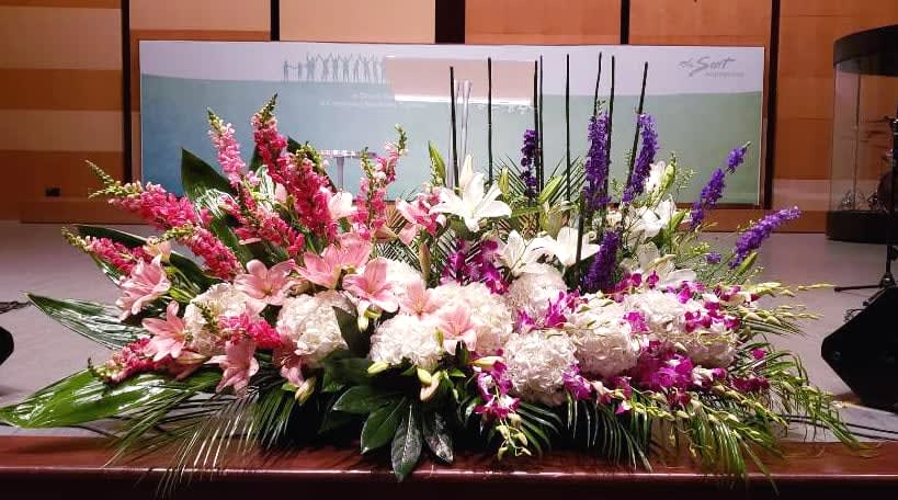 Large low centerpiece of pink lilies, white hydrangeas, and purple flowers along a stage edge