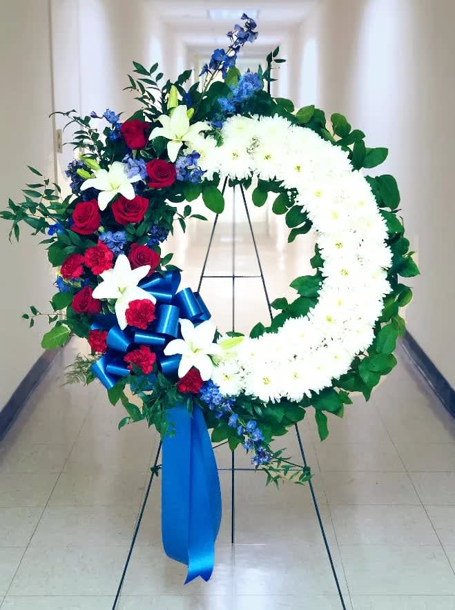 Large floral wreath with white chrysanthemums, red roses, blue flowers, and a blue ribbon
