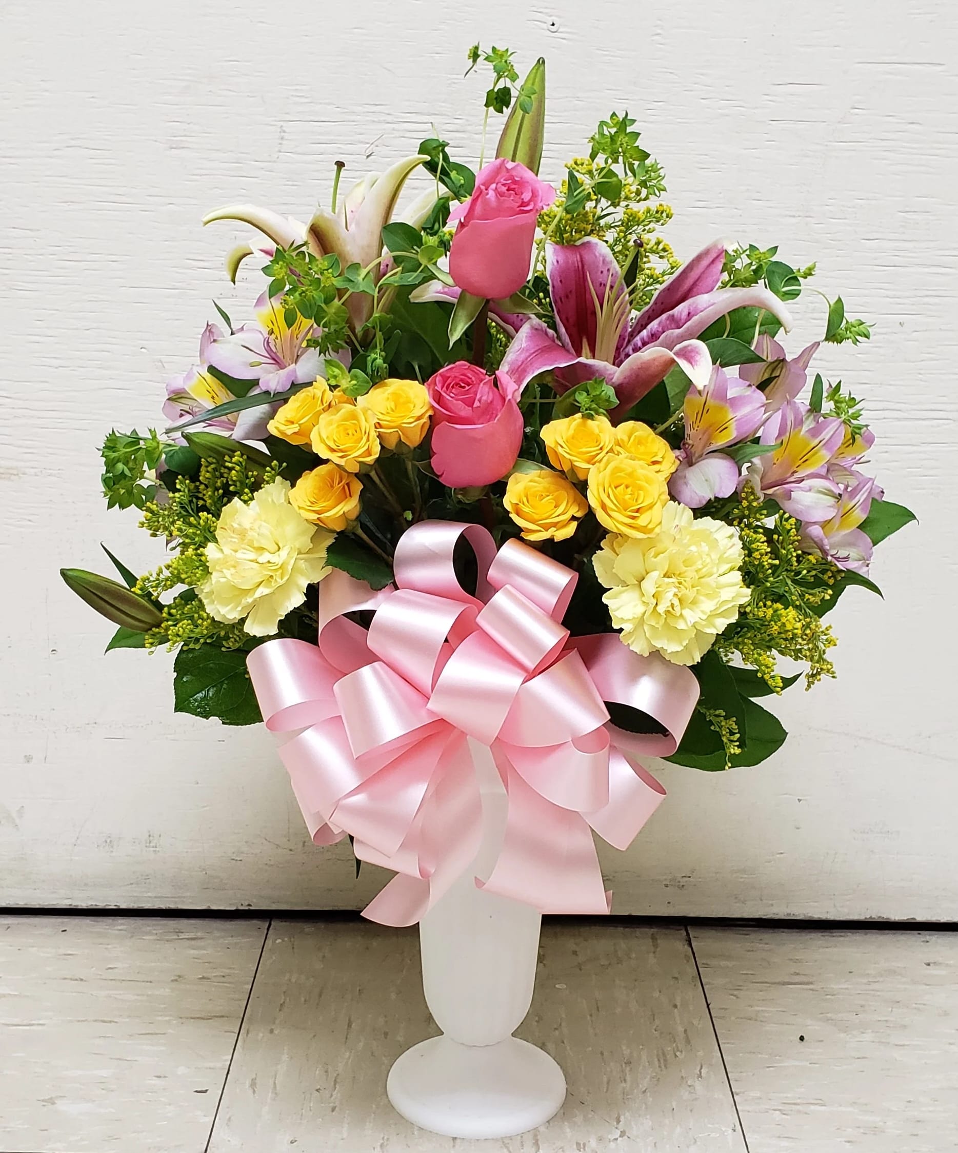 Tall arrangement of pink roses, lilies, yellow spray roses and carnations in a white vase with a pink bow