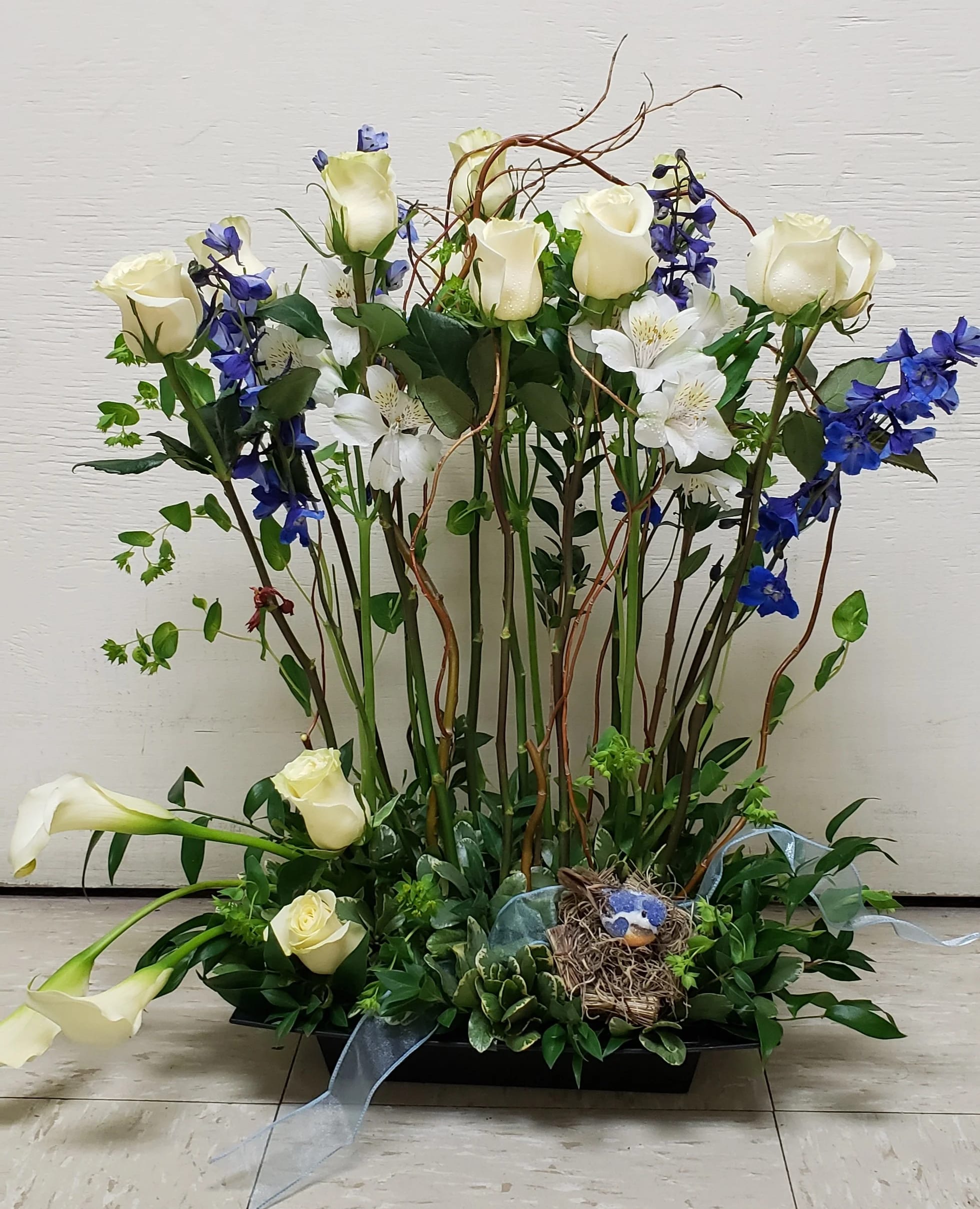 Tall floral arrangement with white roses, blue flowers, and calla lilies in a black tray