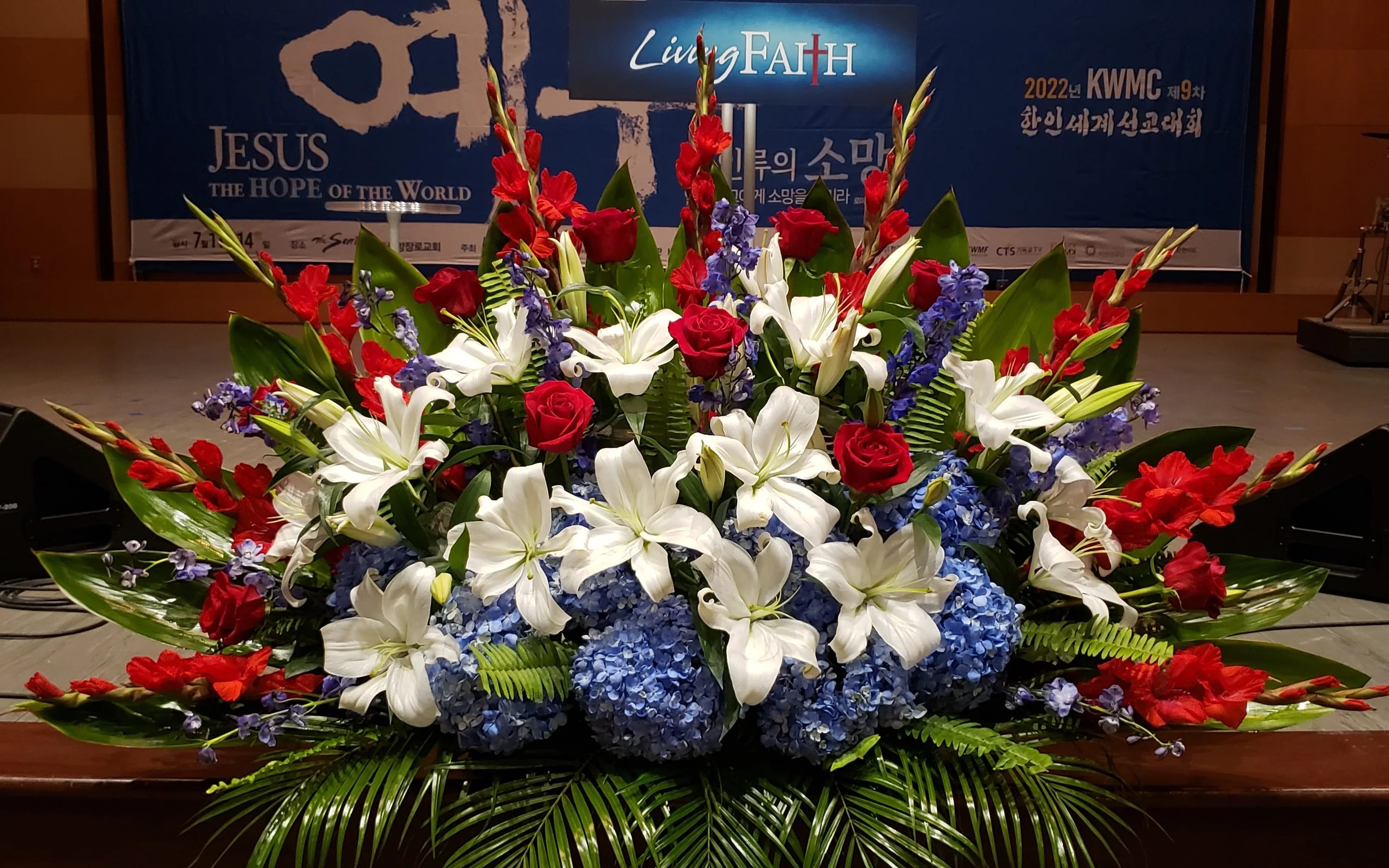 Large red, white, and blue stage arrangement with lilies, roses, hydrangeas, and palm fronds.