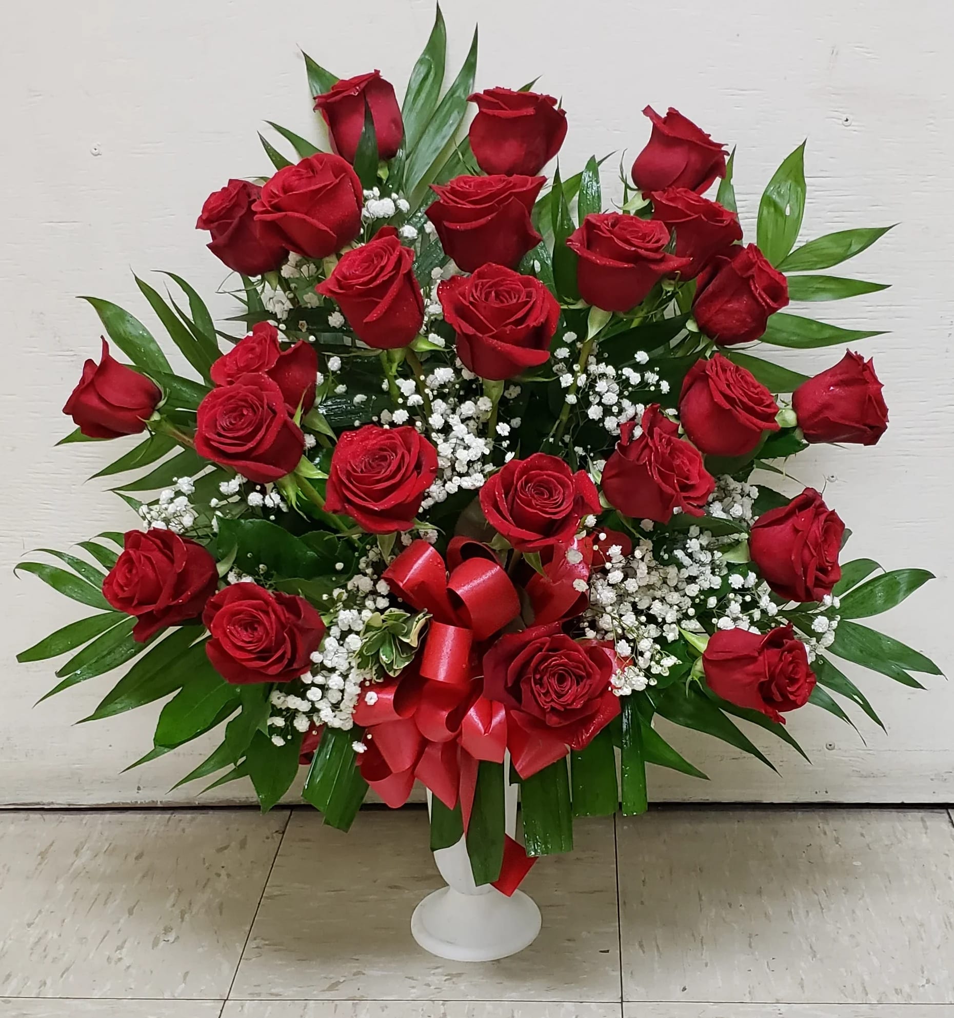 Red roses arranged in a white pedestal vase with baby's breath and a red ribbon