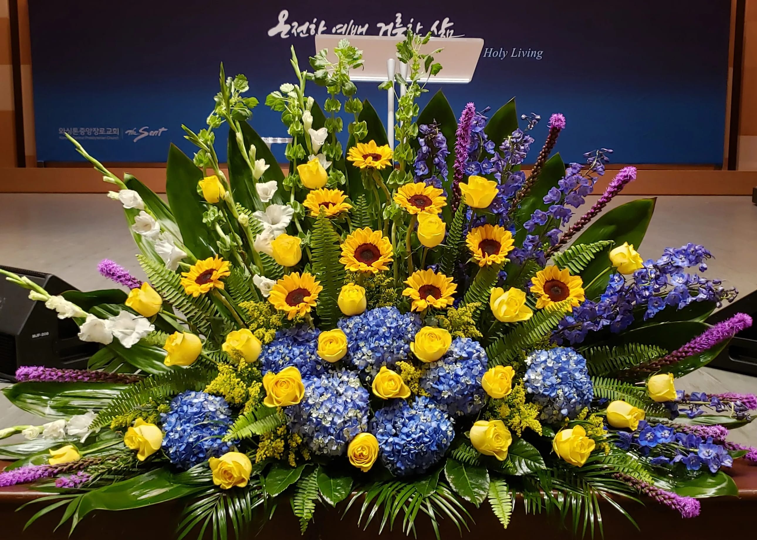 Large altar arrangement of yellow roses, sunflowers, blue hydrangeas, and tall purple accents