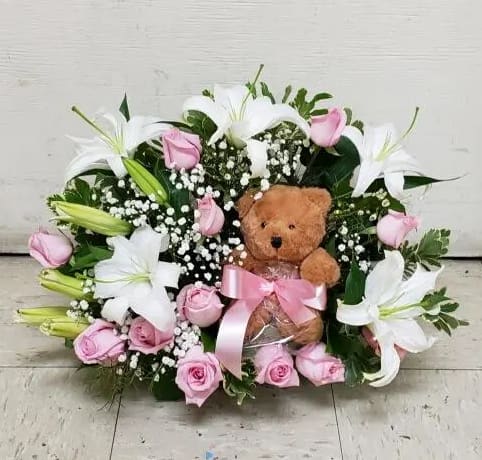 Pink roses and white lilies arranged around a teddy bear with a pink ribbon