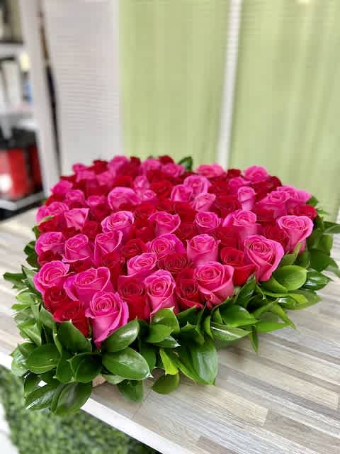 Heart-shaped arrangement of pink and red roses edged with glossy green foliage on a wooden surface