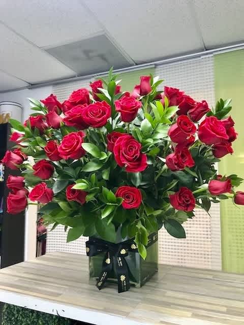Large arrangement of red roses with abundant greenery in a clear glass cube vase tied with a black ribbon.