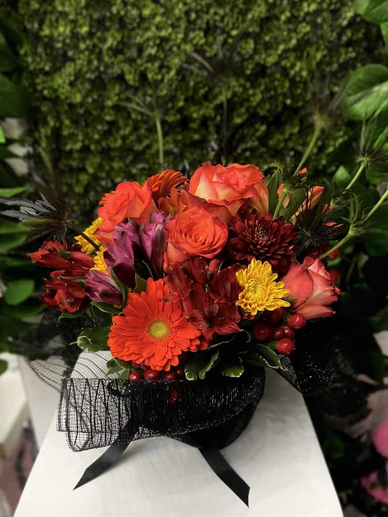 Compact arrangement of red and orange roses, gerbera daisy, and mixed blooms with black mesh wrap