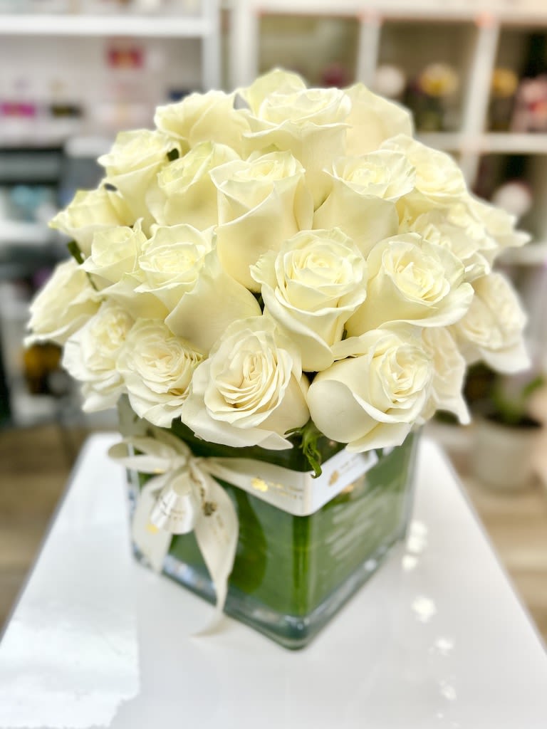 Compact arrangement of cream roses in a clear square glass vase with a ribbon bow.