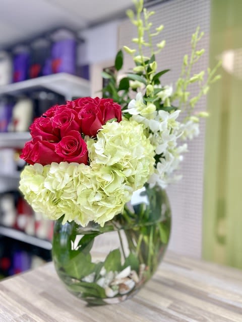Asymmetrical arrangement of red roses, green hydrangeas, and white orchids in a round glass vase with shells.