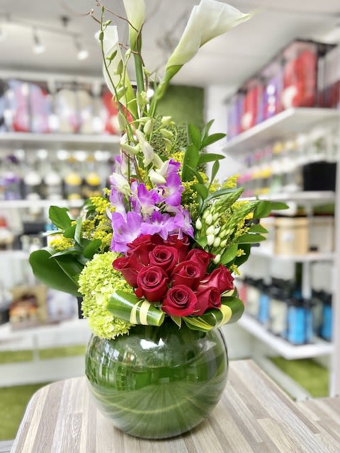 Tall arrangement with white calla lilies, purple orchids, and red roses in a round green-lined glass vase