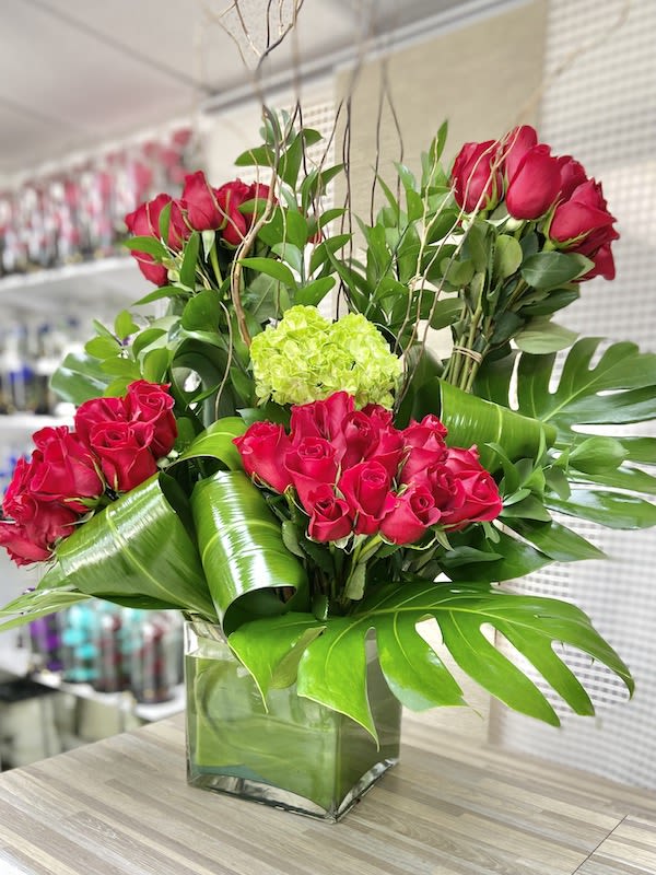 Tall arrangement of red roses with large tropical leaves in a square glass vase
