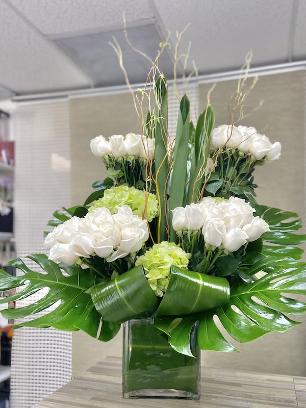 Tall modern arrangement of white roses and green hydrangeas in a square glass vase with large tropical leaves.