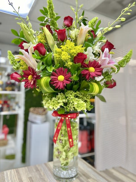 Tall mixed arrangement of red roses, pink lilies, and daisies in a clear cylinder vase with a red ribbon