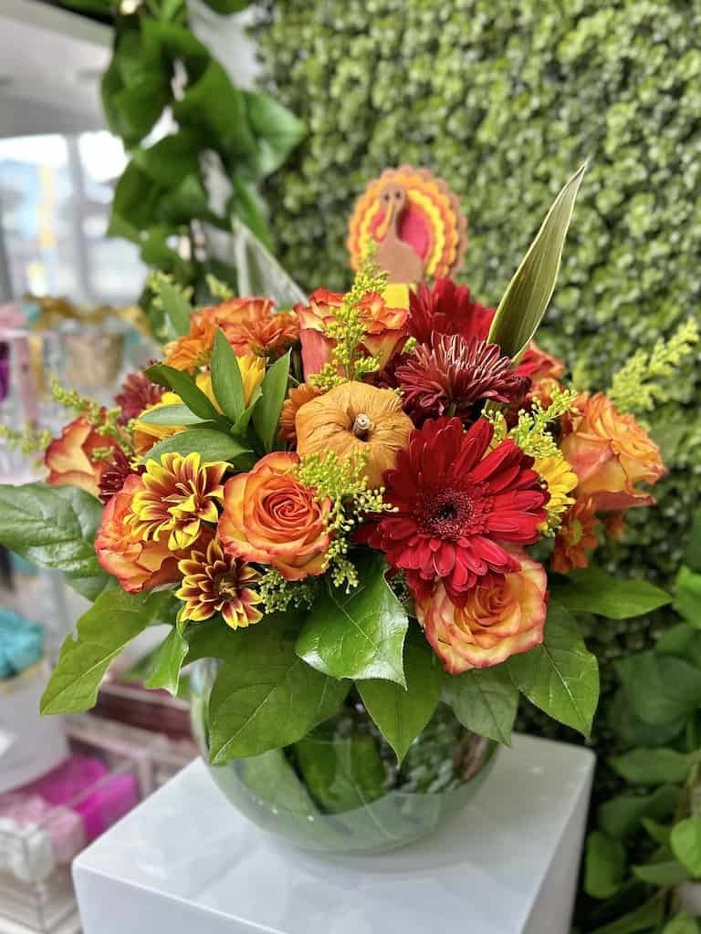 Fall arrangement of orange roses and red gerbera daisies in a round glass vase with a turkey pick