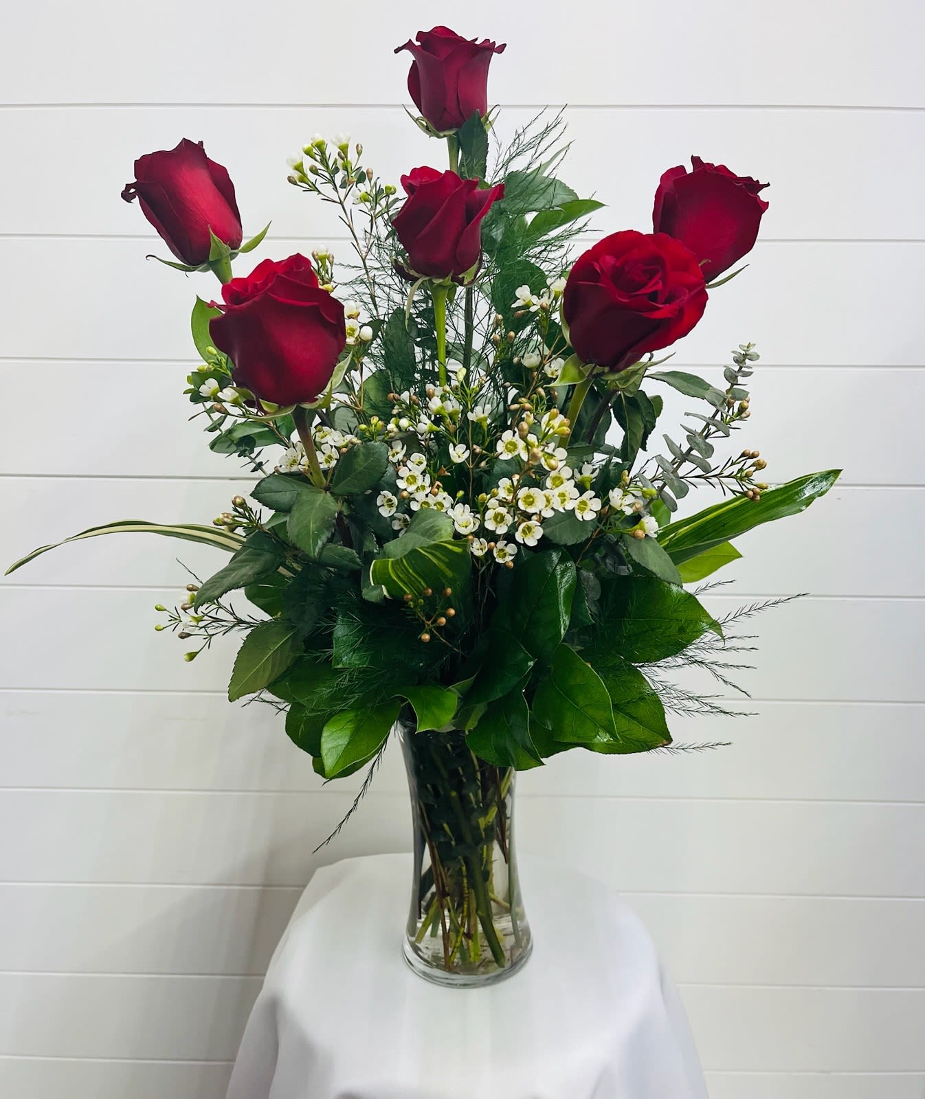Tall arrangement of five red roses with small white blooms in a clear glass vase