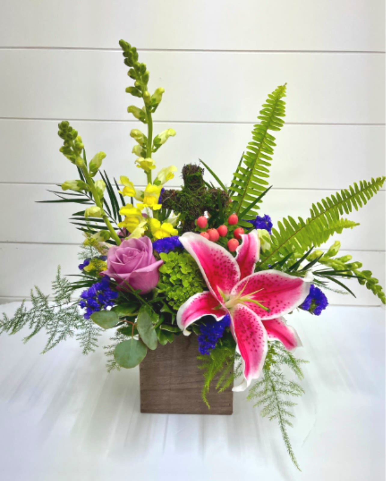 Mixed arrangement with pink lily, purple rose, yellow snapdragons, and ferns in a wooden cube.