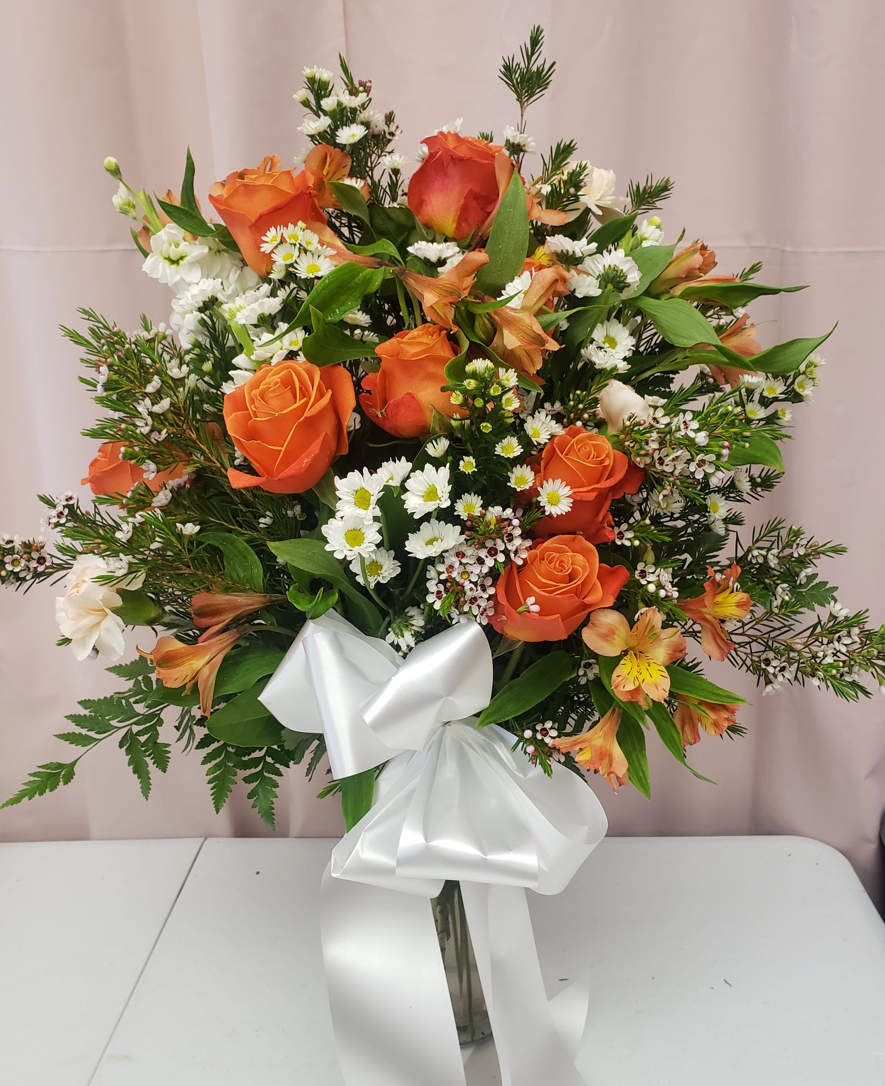 Orange roses and white daisies in a clear vase with a white ribbon
