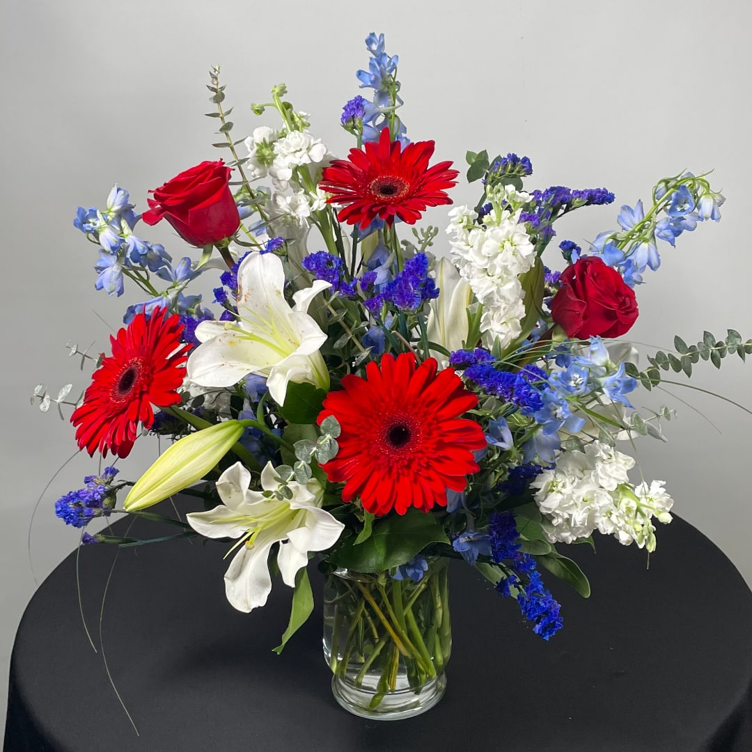 Mixed bouquet with red gerbera daisies, red roses, white lilies, and blue flowers in a glass vase