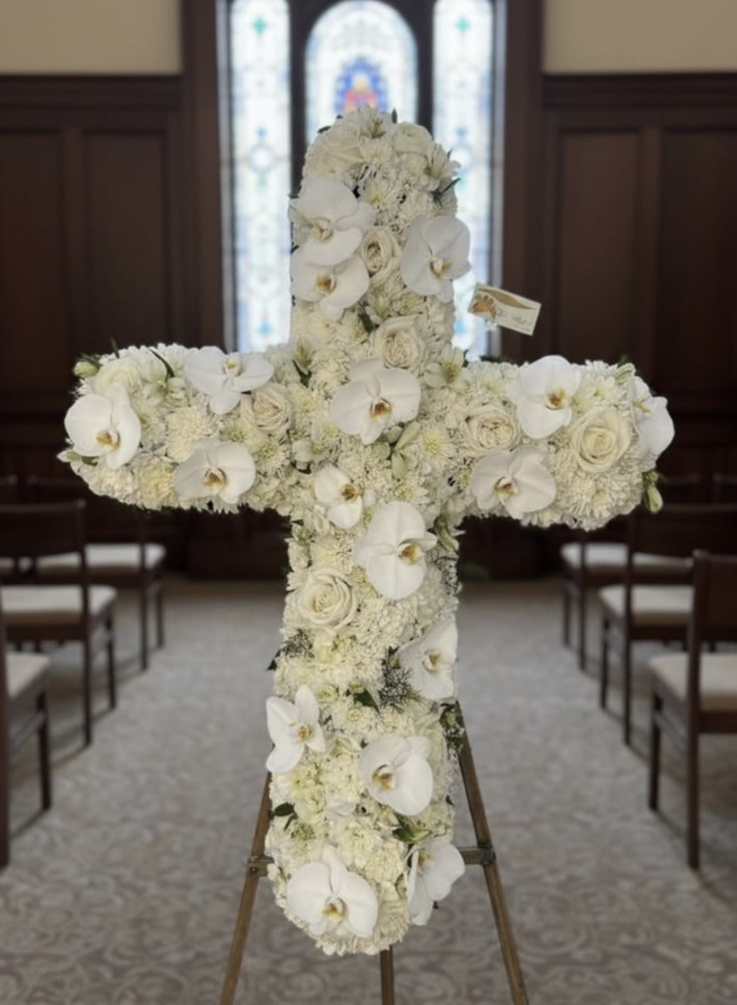 Tall white floral cross with orchids and roses displayed on an easel in a chapel