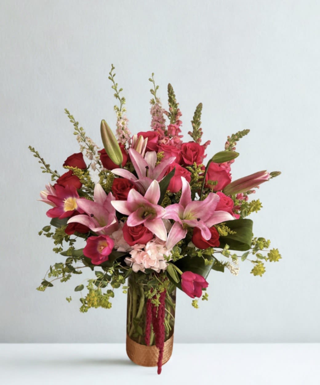 Lush Romance  - A refined mix of red and hot pink roses with soft pink lilies, accented by hot pink tulips, snapdragons, and delicate pink larkspur—arranged with lush greenery in a gold-detailed vase for a bold, elegant statement. The standard version offers the same design in a smaller size, presented in a clear vase.