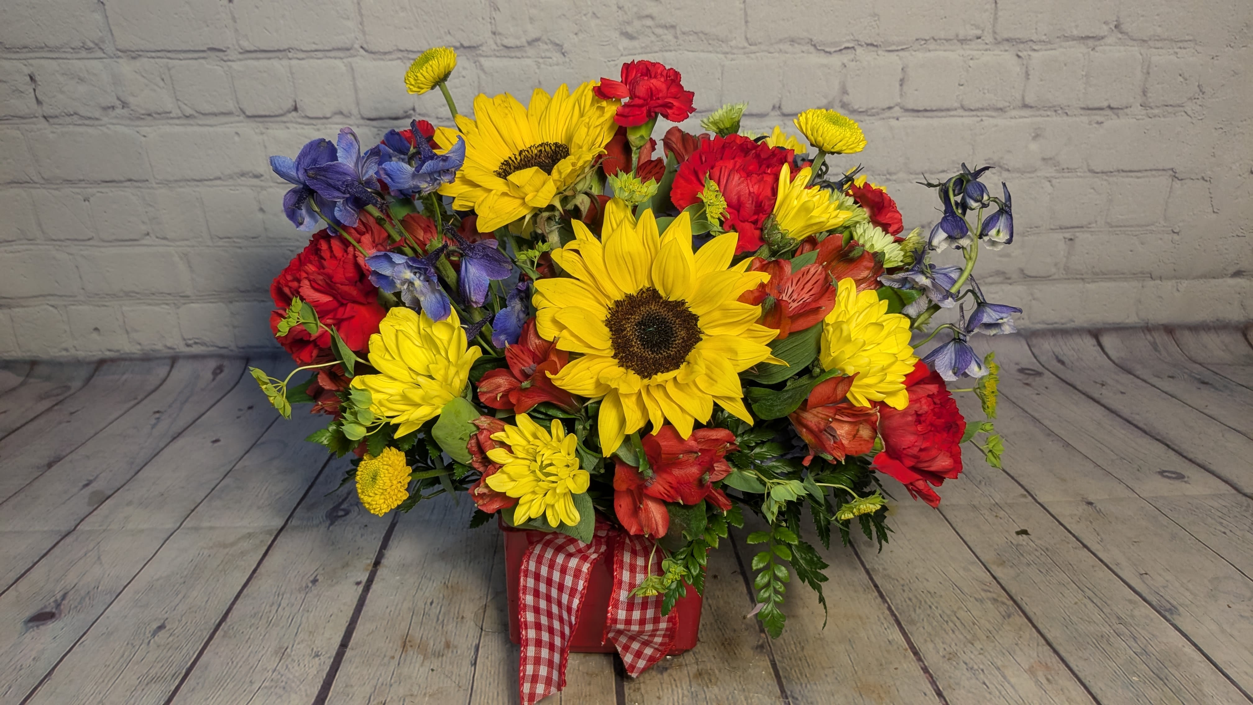 Americana Sunshine - Bursting with color and country charm, our Americana Sunshine bouquet is a cheerful nod to classic summer days. Designed in a bold red cube and tied with a red gingham bow, this arrangement overflows with golden sunflowers, vibrant blue delphinium, sunny yellow daisies, and rich red alstroemerias and carnations. Full of rustic beauty and bright energy, it’s the perfect gift for celebrations, summer gatherings, or anyone who could use a dose of sunshine.