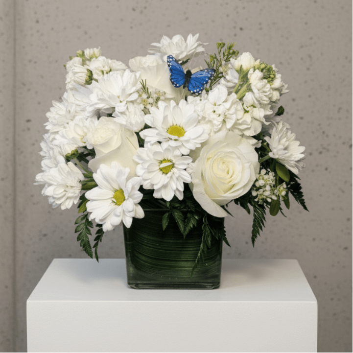 White flowers arranged in a square glass vase with a blue butterfly accent.