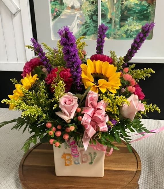 Colorful baby-themed flower arrangement in a white vase with pink ribbon