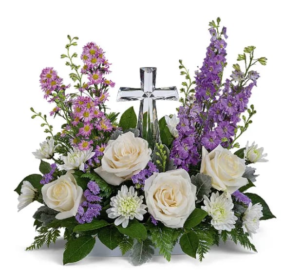 White roses and purple flowers arranged around a clear glass cross.