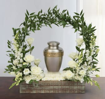 White roses and greenery arranged around a metal urn in a wooden box