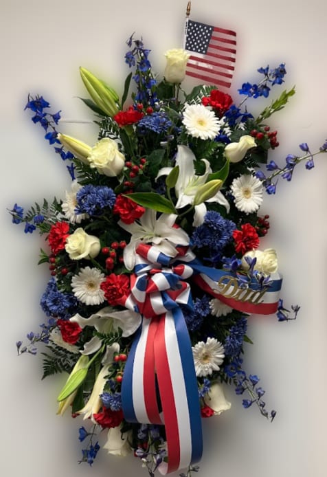 Patriotic Tribute Spray - This red, white, and blue standing spray is a patriotic and meaningful tribute, designed to honor a loved one with dignity and pride. Featuring a beautiful blend of fresh flowers in classic red, white, and blue tones, this arrangement creates a bold and respectful display.  Expertly arranged on a standing easel, it is suitable for funeral services or memorials, offering a heartfelt expression of remembrance and respect.  If you would like a script included on the ribbon, please note your request in the florist note section when placing your order.  For delivery, please enter the name of the deceased in the recipient field and include the correct date of the service to ensure proper timing and placement. 