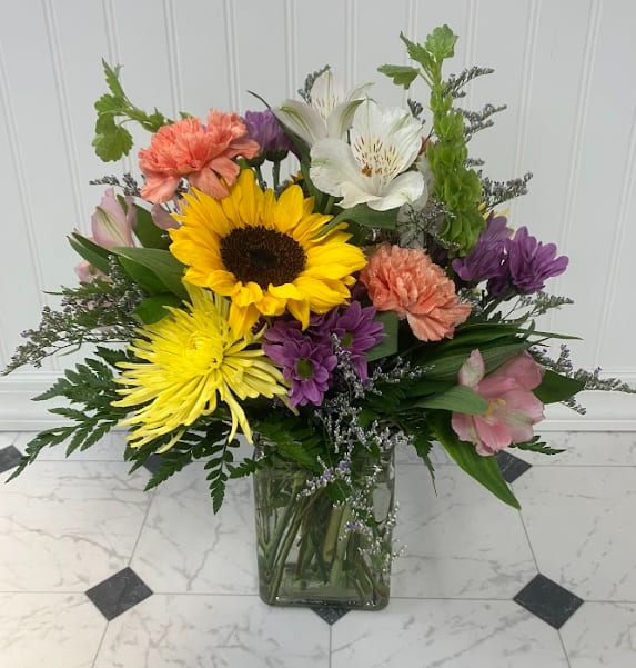 Sunlit Meadow - Inspired by the beauty of a wildflower meadow, Sunlit Meadow features radiant sunflowers paired with orange, yellow, purple, and white blooms for a warm and colorful display. Accents of Bells of Ireland add height and texture, creating a fresh arrangement full of sunshine and natural beauty.