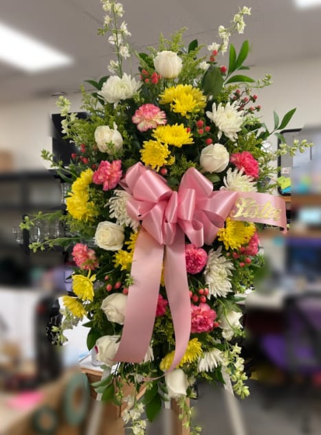 Standing spray of pink, yellow, and white flowers with a large pink bow on an easel.