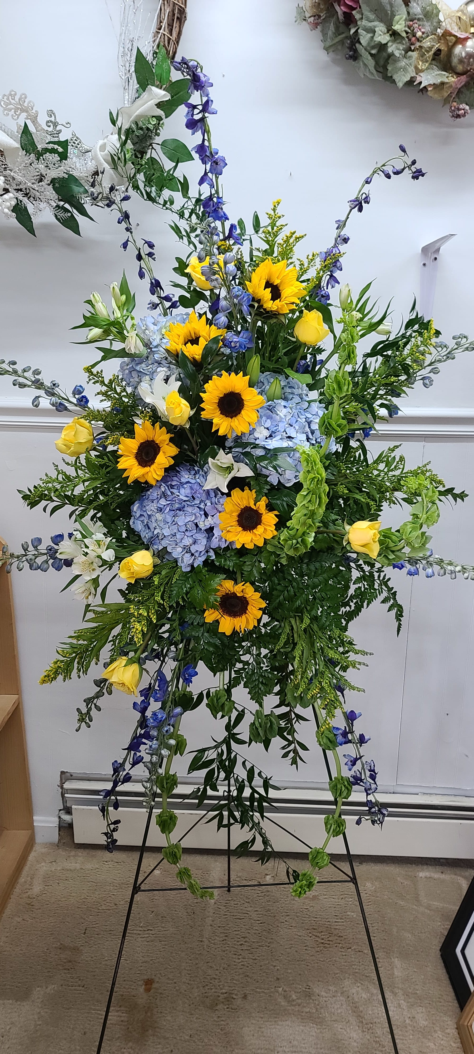 Sunflowers on Blue - Traditional Standing spray of golden sunflowers, yellow roses, blue delphinium, bells of ireland, Hydrangea, and alstromaria complement each other and create an outstanding display. This beautiful, showy, tribute can be sent with or with out a bow. If you would like script on the ribbon, please leave what you would like in the "special instructions" section or ask us to call you. 