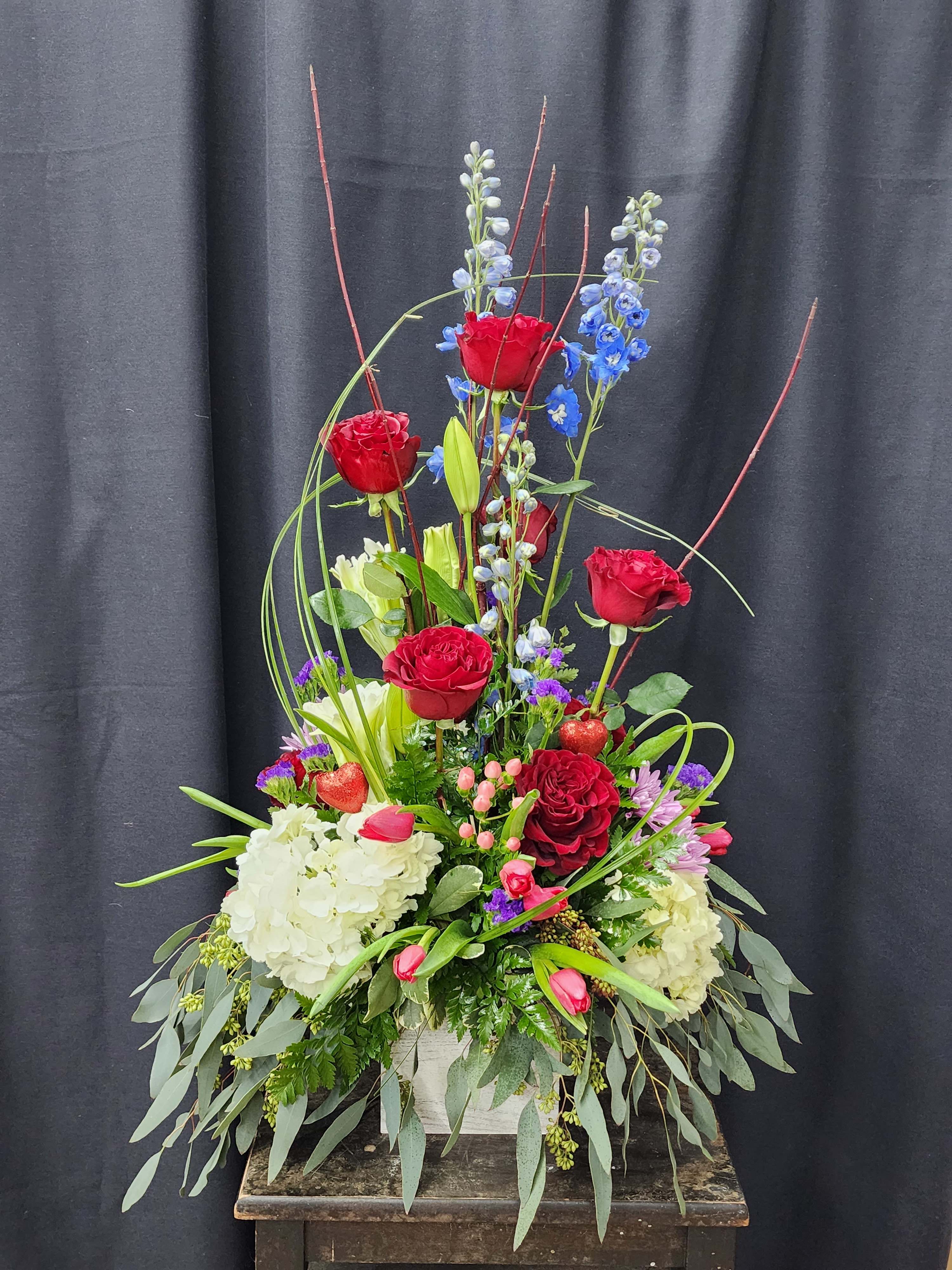 Tall floral arrangement with red roses, blue delphinium, and white hydrangeas