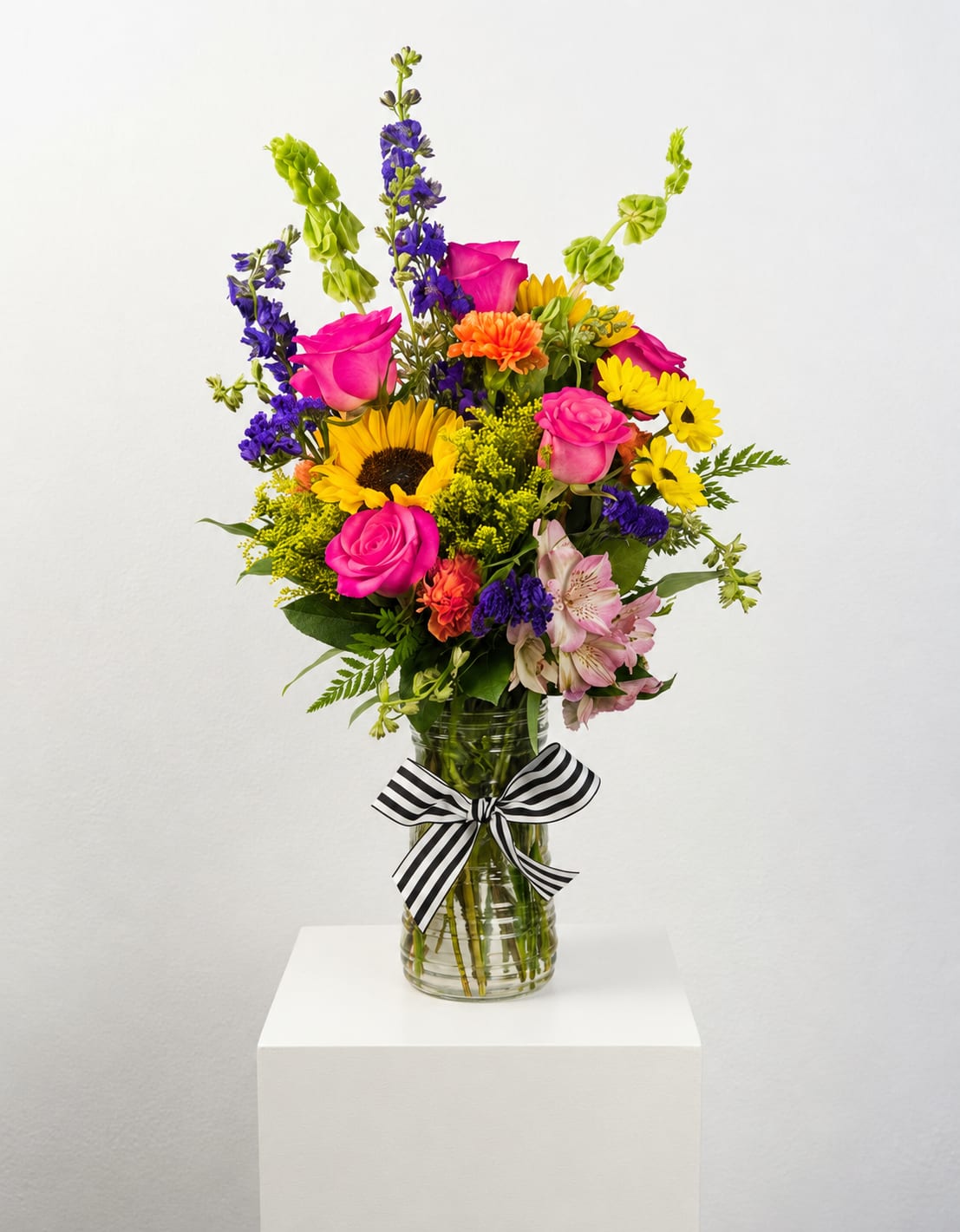Here Comes the Sun - This arrangement full of bright and cheery colors will have you looking ahead to warmer days. Here Comes the Sun has sunflowers, bells of Ireland, blue delphinium, alstroemeria, and roses as well as seasonal flowers and greens that will have you walking along Abbey Road and humming this classic Beatles song.   Please Note: Standard Delivery for this product occurs in the afternoon, between 2:30pm and 7pm. If you need it by a certain time, there is an option to select a timed window for scheduled deliveries.