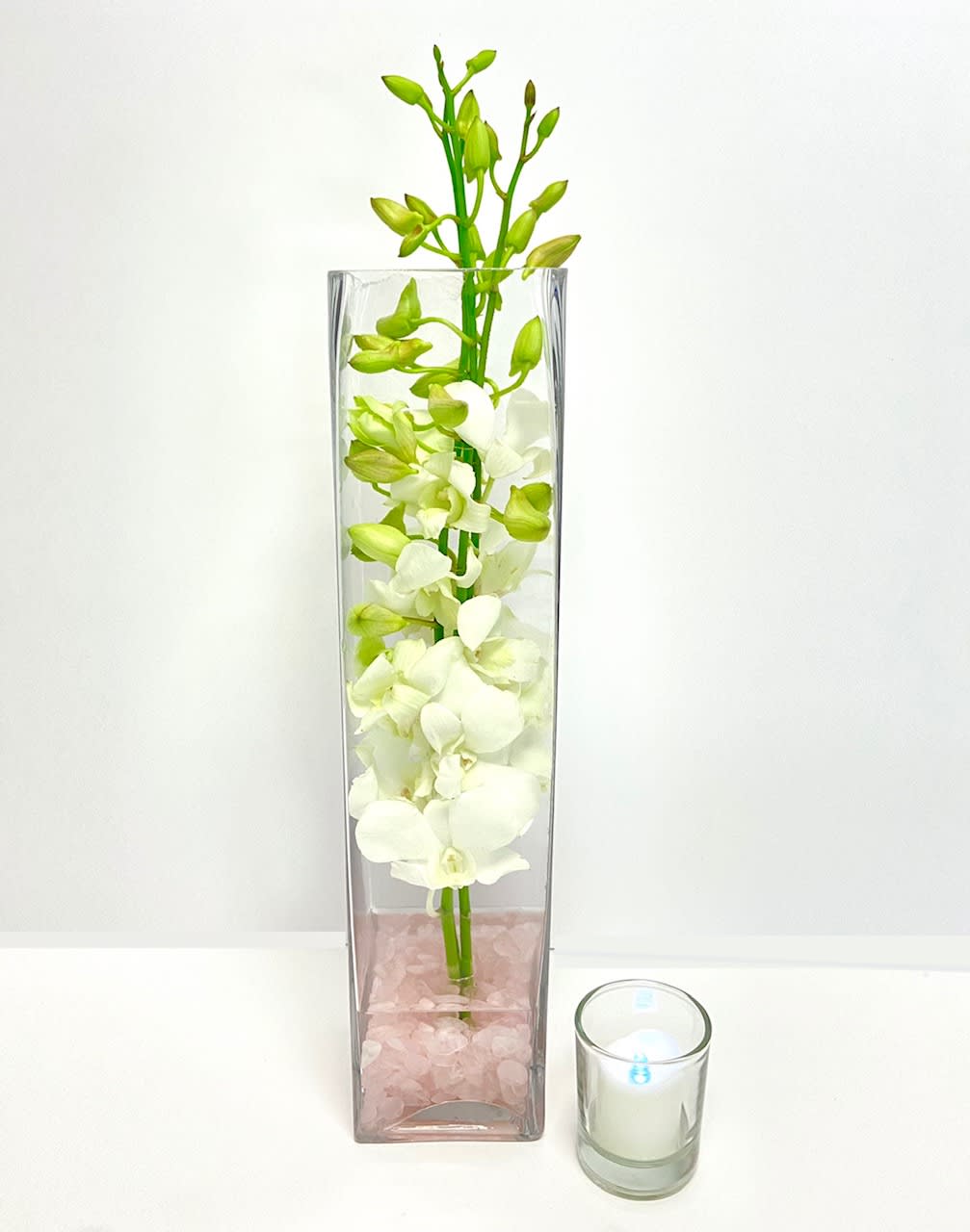 White orchid stems in a tall glass vase with pink stones and a small candle beside it