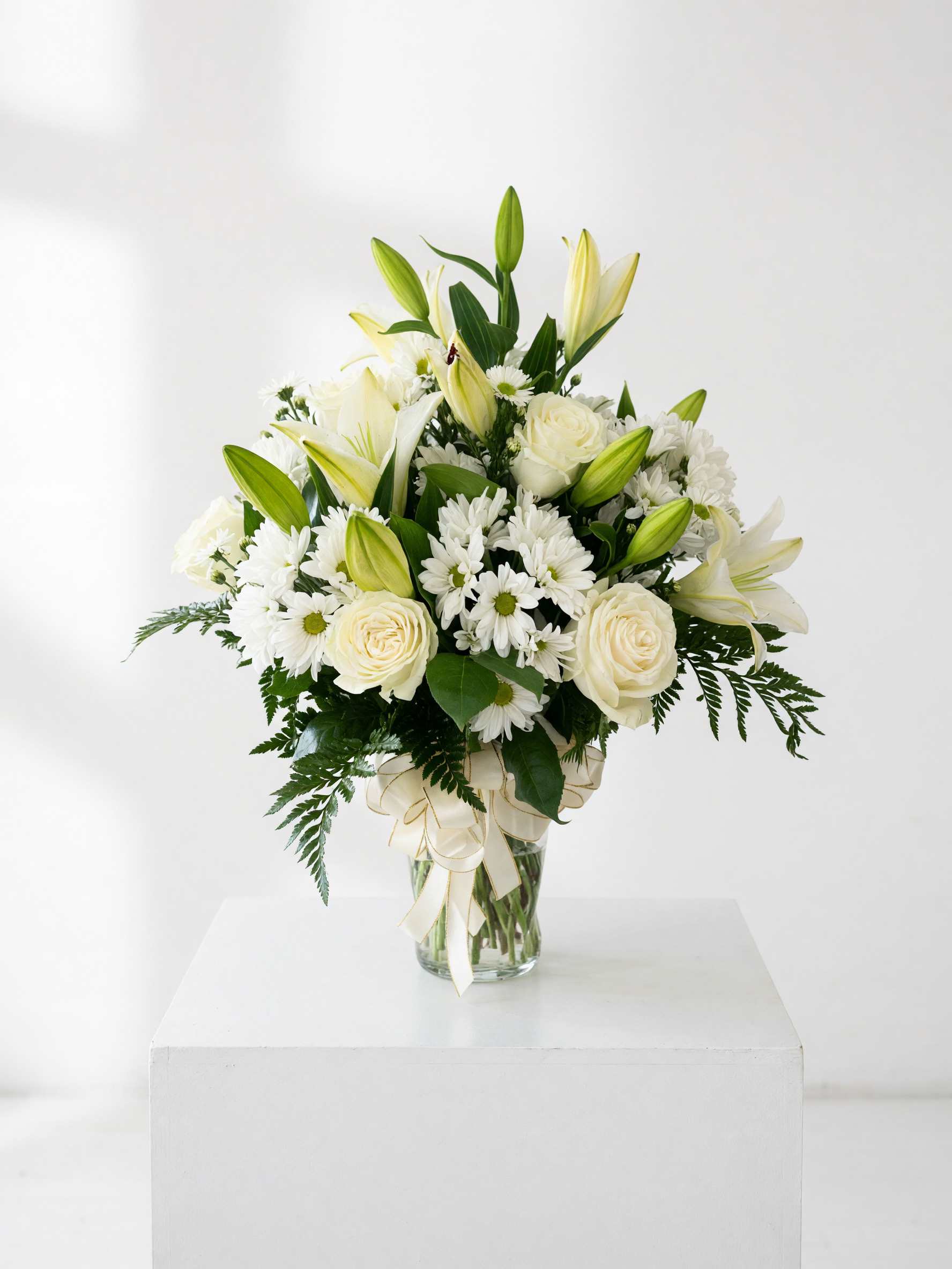 White lilies, roses, and daisies arranged in a glass vase with a ribbon
