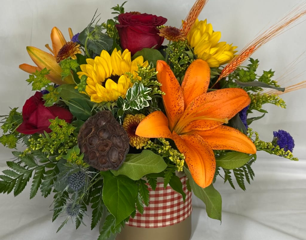 Mixed bouquet with orange lilies, red roses, and yellow sunflowers in a gingham-wrapped container.