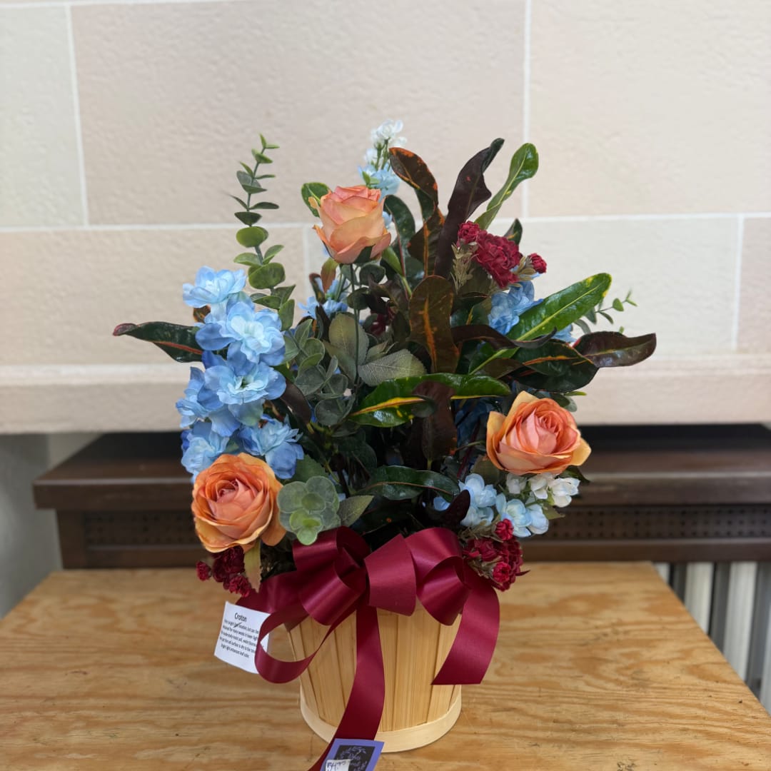 Peach roses and blue flowers arranged in a wooden basket with a burgundy ribbon.