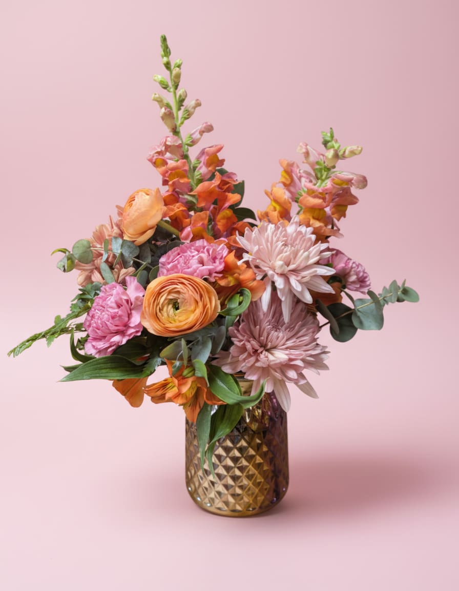 Coral Kiss - A romantic celebration of spring in full bloom, Coral Kiss is arranged in a luminous iridescent vase and features coral ranunculus, pink cremones, blush carnations, orange alstroemeria, and orange snapdragons. Softly layered and gracefully balanced, this arrangement exudes elegance and warmth—perfect for expressing love, joy, or heartfelt sentiment.  All flowers, floral colors, and containers are subject to change due to availability. 