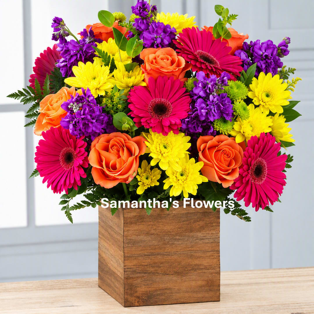Bright mixed bouquet in a wooden cube vase with pink, orange, yellow, and purple flowers.