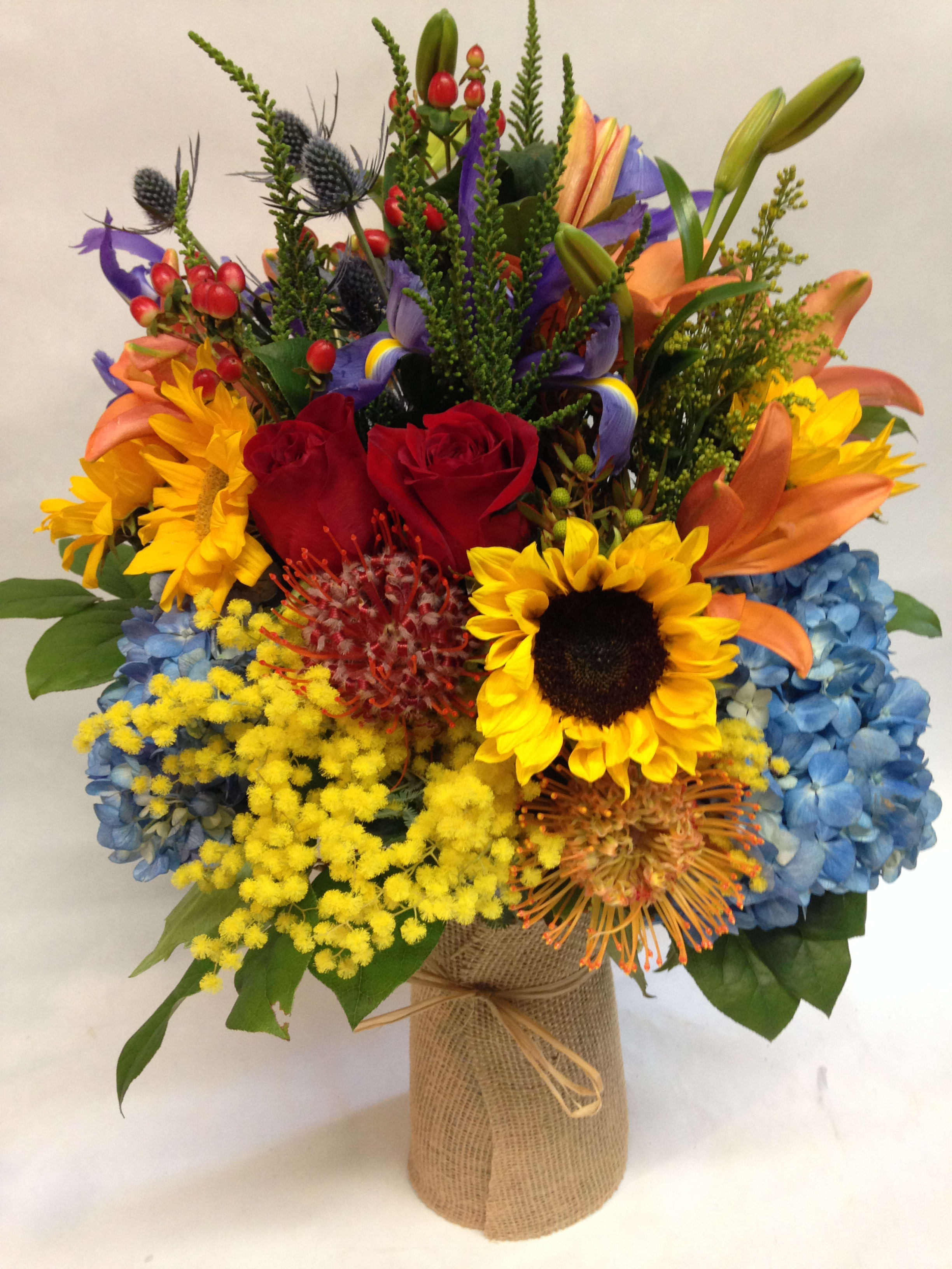 Over the Rainbow - This bright and cheerful arrangement is complete with hydrangea, sunflowers, roses, pin-cushion proteas, asiatic lillies, sea thistles and irises.  (Some flowers may be subject to change depending on the season.  We will contact you with available flower substitutions; please contact us with specific questions if you are concerned with flower availability, thanks!) This arrangement comes with a burlap wrapped vase.  Arrangement featured: $325.00 (Regular Price)
