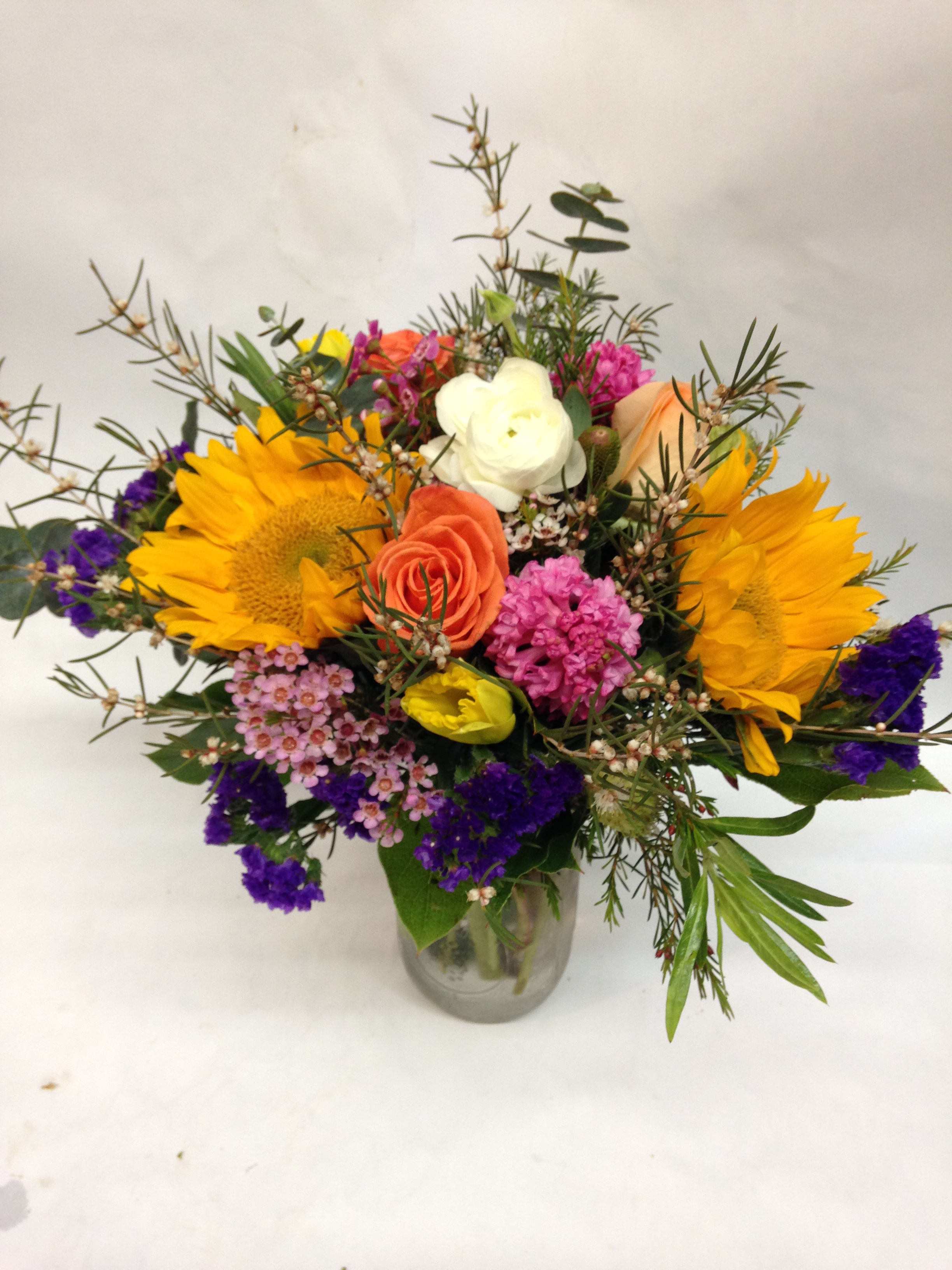 Mixed bouquet with sunflowers, roses, and purple filler in a glass vase