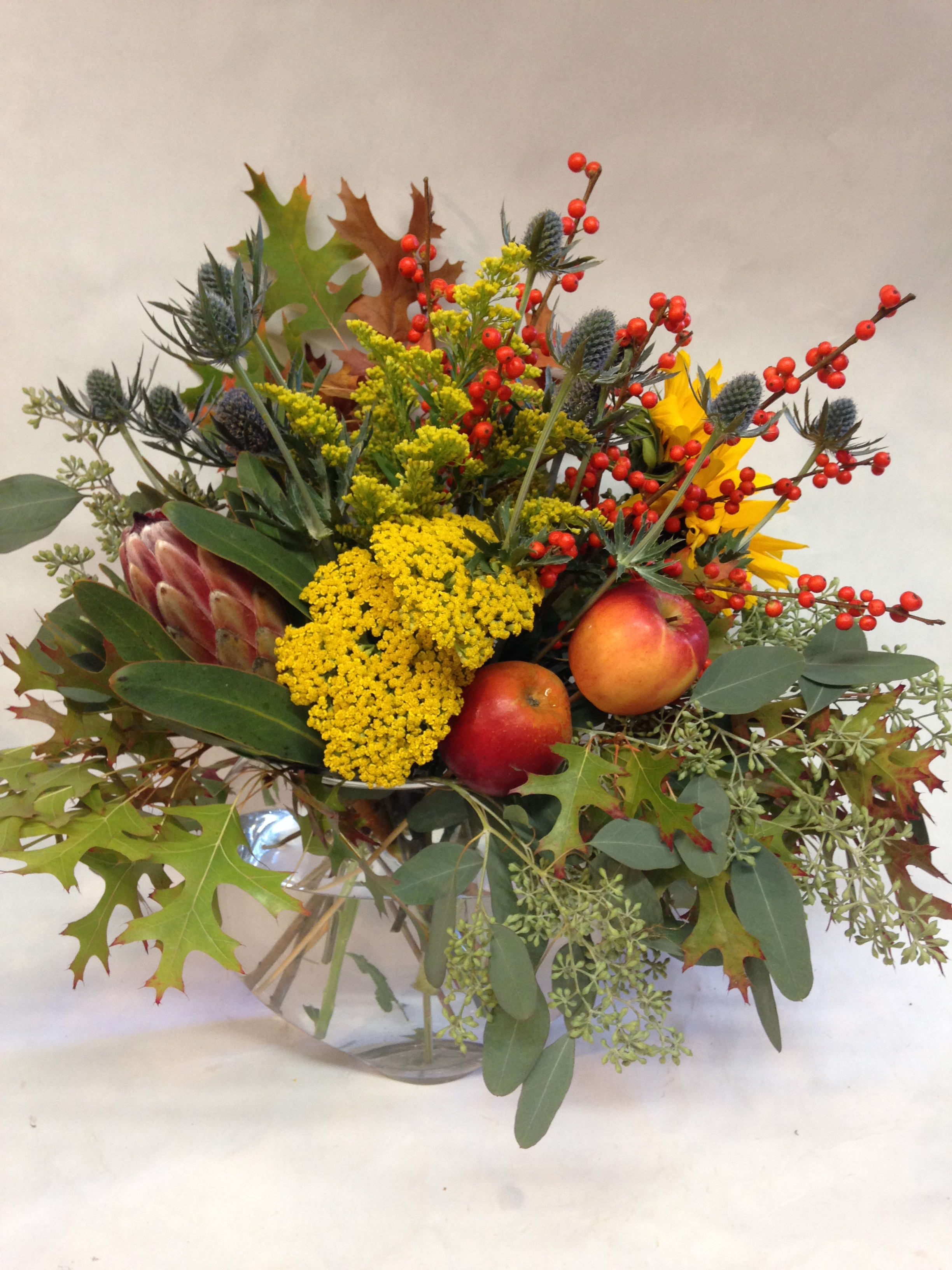 Autumn bouquet with yellow flowers, red berries, and apples in a glass vase