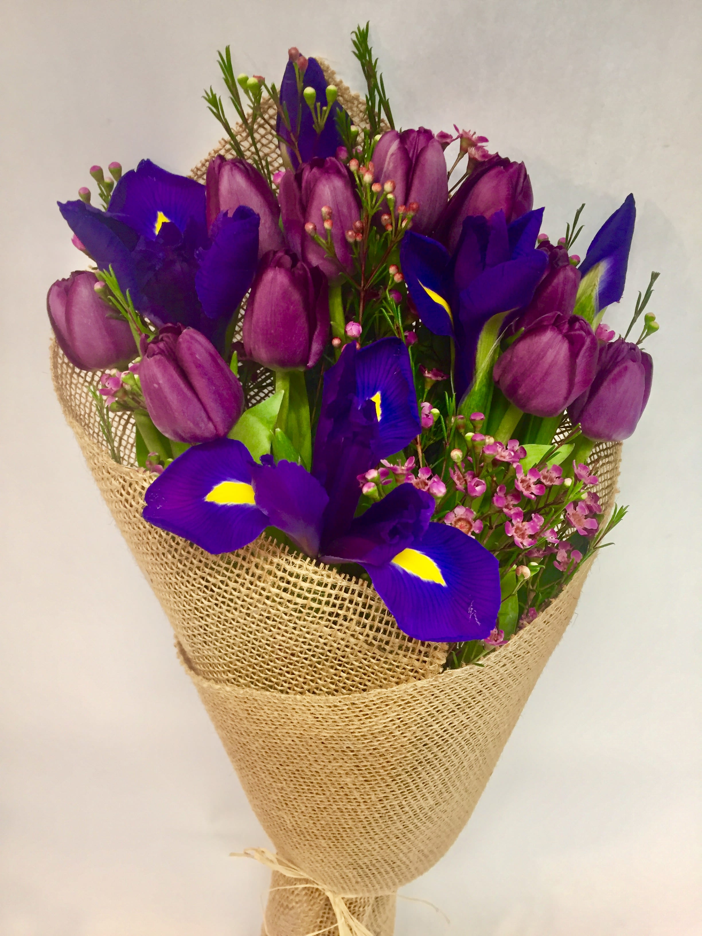 Midnight Iris &amp; Amethyst Tulip Bouquet - A striking hand-tied bouquet featuring rich purple tulips and vibrant royal-blue irises accented with delicate pink waxflowers and fresh greenery. Wrapped in rustic natural burlap, this elegant arrangement combines bold jewel tones with a touch of countryside charm. The dramatic contrast of deep violet blooms and bright yellow iris highlights creates a sophisticated and memorable gift, perfect for birthdays, anniversaries, celebrations, or simply brightening someone’s day.